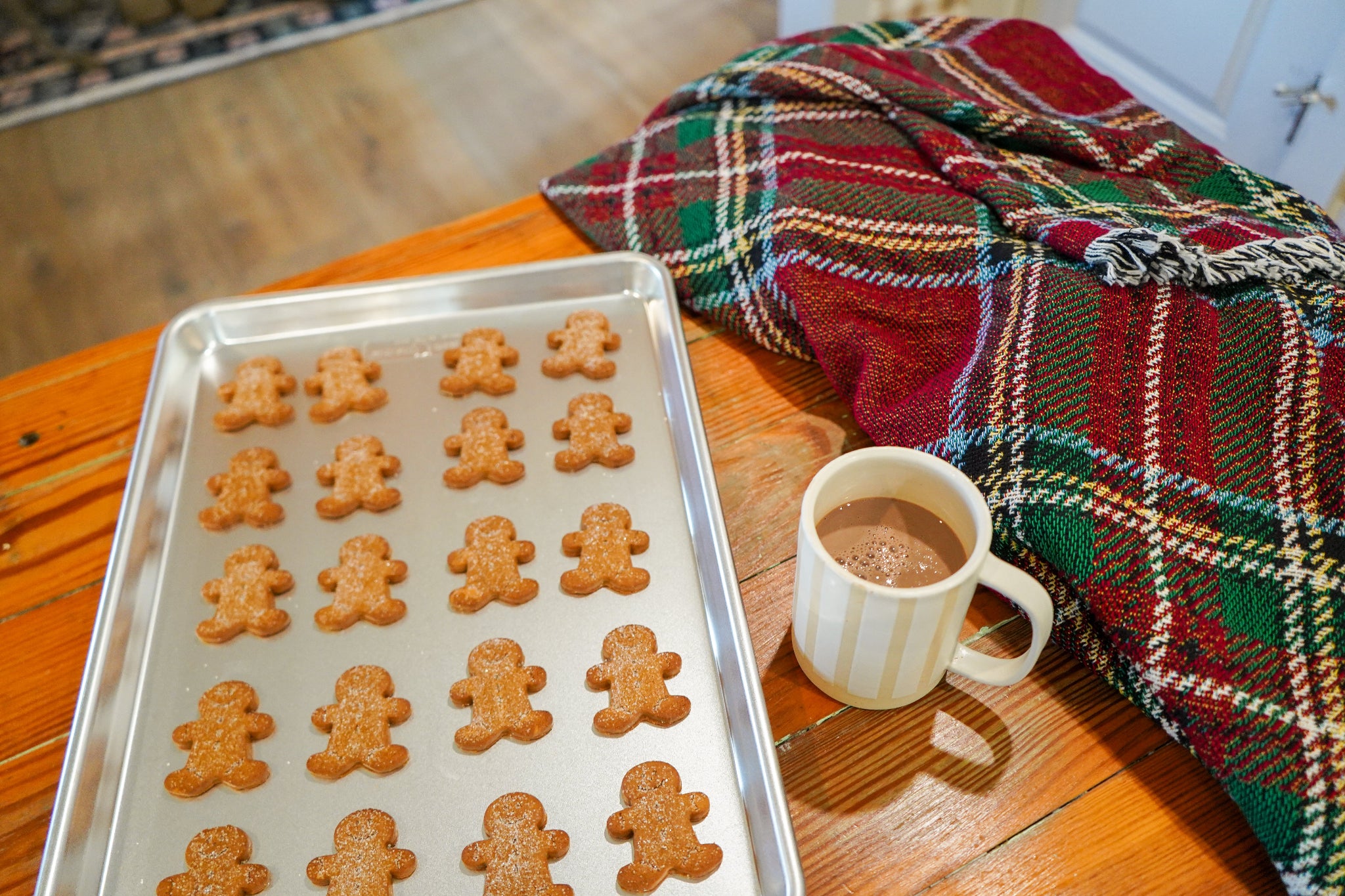 Sheet of gingerbread men cookies on a baking tray with a mug of hot chocolate and plaid blanket in the background.
