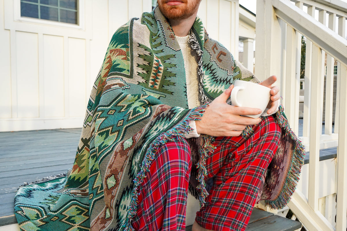 Man sitting on a porch with a patterned blanket and plaid pants, holding a mug.